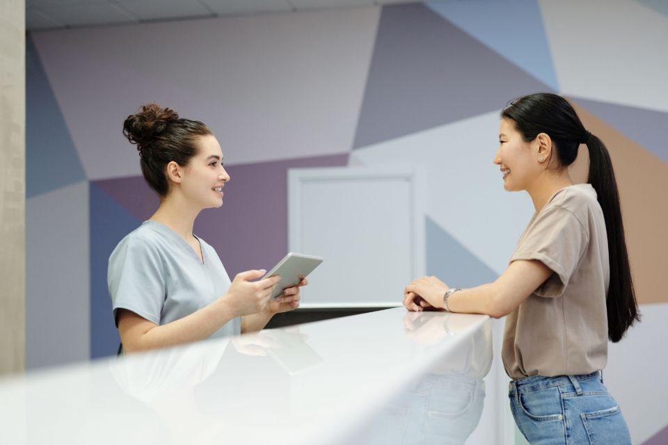 A receptionist holding a tablet talks with a patient across the reception counter.