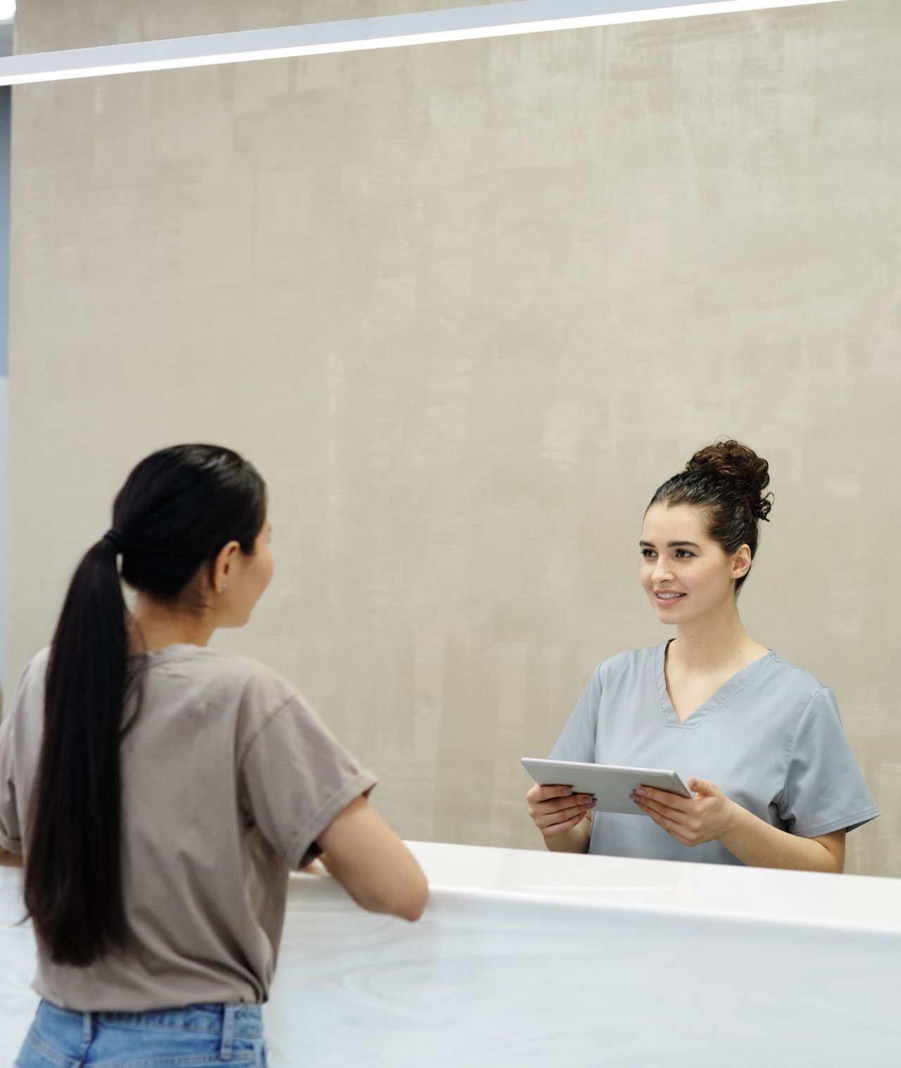 A dental receptionist in scrubs smiles as she helps a patient at a bright, modern front desk.